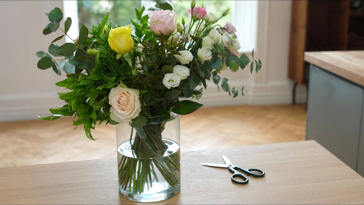 A fresh mixed bouquet in a clear glass vase of water on a light wooden kitchen table by a window, with floristry scissors beside it